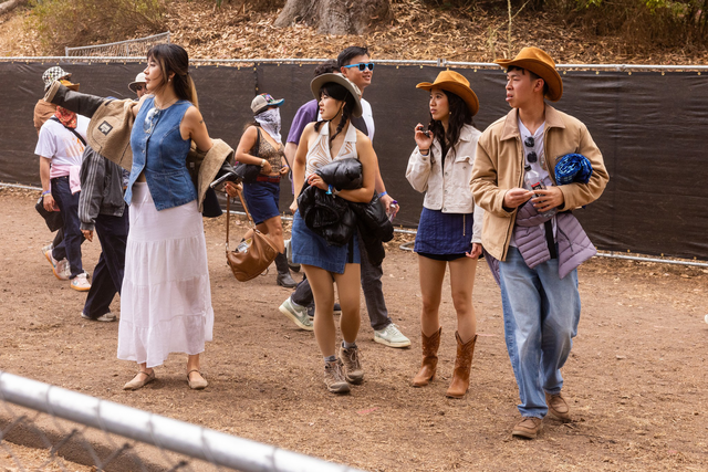 A group of people walk outdoors on dirt, some wearing cowboy hats and boots, with trees and a dark fence in the background.